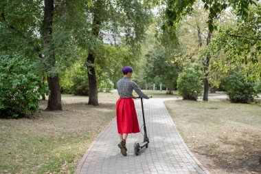 Mor saçlı genç bir kadın parkta elektrikli scooter kullanıyor. Ekose gömlek, uzun kırmızı etek ve papyon bir tıraş tapınak ile şık bir kız modern bir cihaz üzerinde şehir etrafında sürme. 