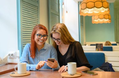 İki heyecanlı genç kız cep telefonu kullanıyor, bir kafede oturuyor ve parmakla işaret ediyor. Gözlüklü kızıl saçlı kadın akıllı telefon kız arkadaşının komik fotoğraflarını gösteriyor. Kahve içerler..