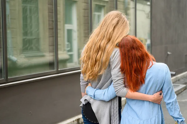 Dos amigos caminan por la calle y se abrazan suavemente alrededor de la cintura. La rubia de la chaqueta y el vestido de mujer pelirroja se inclinaron la cabeza. Vista desde atrás — Foto de Stock