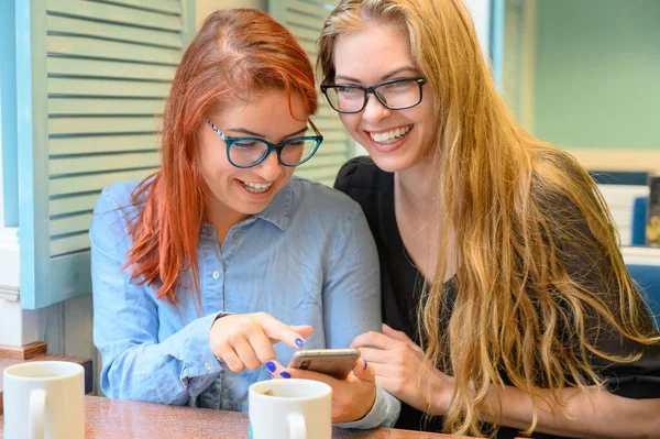 İki heyecanlı genç kız cep telefonu kullanıyor, bir kafede oturuyor ve parmakla işaret ediyor. Gözlüklü kızıl saçlı kadın akıllı telefon kız arkadaşının komik fotoğraflarını gösteriyor. Kahve içerler.. — Stok fotoğraf