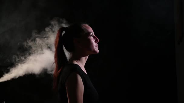 A sad brunette is in a cloud of smoke. Portrait of a woman smoking a vape behind her, soaring. Black background. Passive smoking. Bad habits. — Stock Video