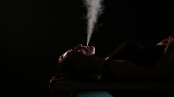 La brune fume une cigarette électronique sur fond noir, dégage une épaisse fumée de sa bouche. Portrait d'une femme fumant un singe, planant .