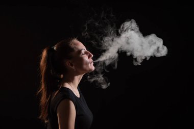 Esmer, siyah arka planda elektronik bir sigara içiyor ve ağzından yoğun bir duman salıyor. Bir kadın portresi sigara içiyor, havada süzülüyor..