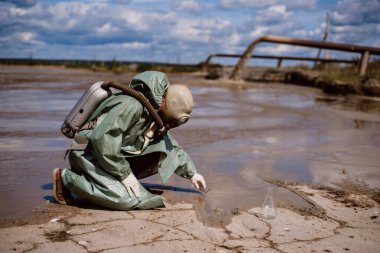 Bir bilim adamı kimyasal atık salınımından sonra su örneği alır. Solunum cihazına bağlı bir adam ve radyasyondan korunan yeşil bir giysi. Su analiz uzmanları. Maske. Ekolojik felaket.