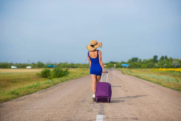 Een vrouw met een hoed en een grote tas loopt langs de weg. Meisje in een blauwe strakke jurk met een paarse koffer op de baan. Brunette met een sportfiguur op de weg. — Stockfoto