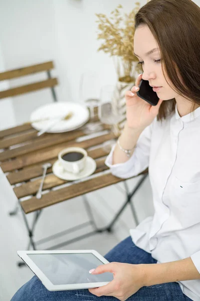 흰색 셔츠와 청바지를 입은 한 여성 이 탁자를 손에 들고 카페에 앉아 있다. 점심 시간에 근무하는 여성 사무실 직원 이 전화로 이야기하는 것이었습니다. 식당에서 커피를 마시는 사업 모임의 지배인. — 스톡 사진