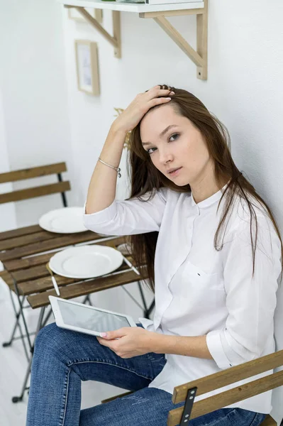 Una joven con una camisa blanca y jeans se sienta en un café en una mesa con una tableta en las manos. Trabajadora de oficina en la hora del almuerzo. Gerente en una reunión de negocios en un restaurante . —  Fotos de Stock