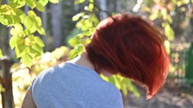 Portre, gözlüklü, gri tişörtlü, kızıl saçlı güzel bir kadın parkta yürüyor ve flört ediyor. Mutlu bir kız döndü ve sıcak bir sonbahar gününde saçlarını salladı. Yaprak dökümü.