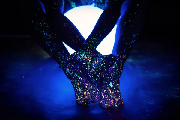 Una donna dipinta con polvere fluorescente tiene una lampada sferica. Primo piano delle gambe di una mano ragazze con spray corpo colorato. Luce notturna rotonda al neon . — Foto Stock