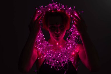 Cadı makyajlı ve burnunda küpe olan bir kadın. Kız yüzünde titreyen pembe ışıklardan oluşan bir çelenk tutuyor. Ampuller karanlıkta cadıların yüzünü aydınlatır..