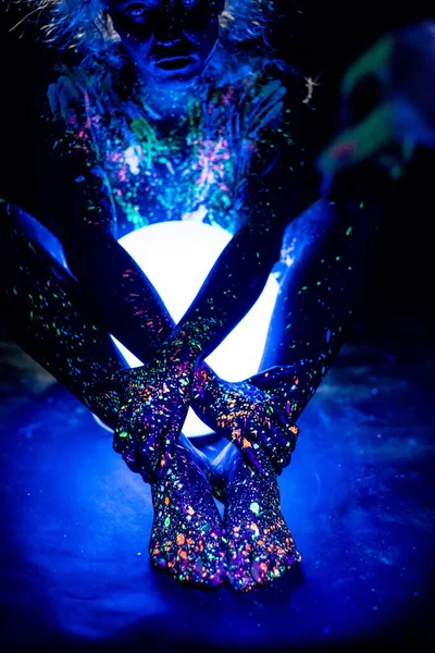 Una donna dipinta con polvere fluorescente tiene una lampada sferica. Primo piano delle gambe di una mano ragazze con spray corpo colorato. Luce notturna rotonda al neon . — Foto Stock