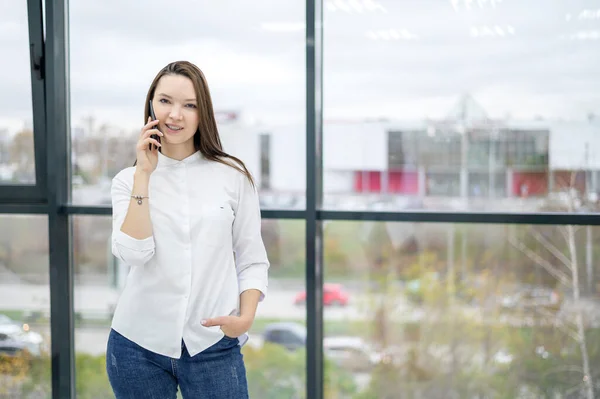 Una mujer con una camisa blanca está junto a la ventana y habla por teléfono. Chica europea está llamando a un teléfono inteligente y sonriendo . —  Fotos de Stock