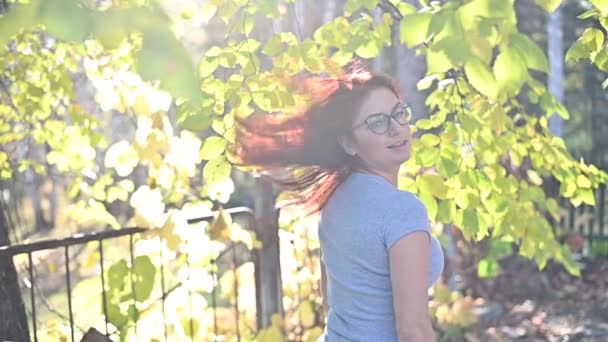 Porträt Schöne rothaarige Frau mit Brille und grauem T-Shirt spaziert durch den Park und flirtet. Ein glückliches Mädchen dreht sich um und wedelt an einem warmen Herbsttag mit ihren Haaren. Laubfall. — Stockvideo