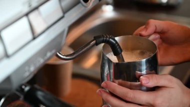 Bir kadın kahve makinesinde kahve hazırlıyor. Ellerin yakın planı. Barista kafede kapuçino için süt ısıtır. Lokantanın mutfağı..
