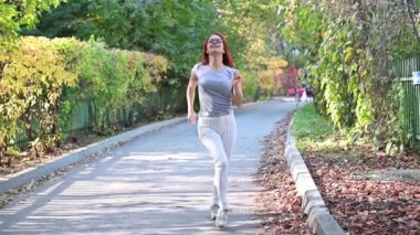 Güzel kızıl saçlı, gözlüklü bir kadın sokakta yürüyor ve dans ediyor. Gri tişörtlü ve çizgili taytlı mutlu kız sonbahar parkında yürüyor ve eğleniyor. Yavaş çekim.