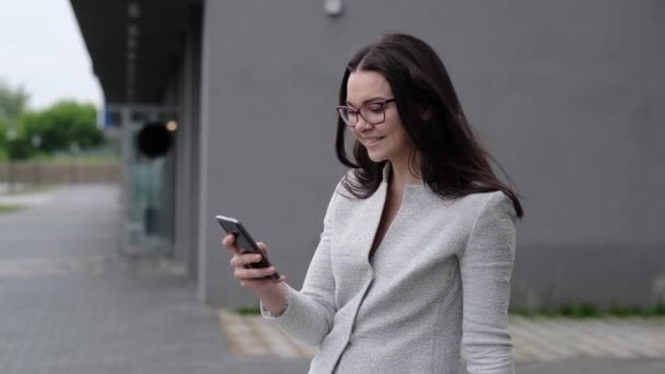 Una joven con un traje de negocios y lee un mensaje por teléfono y sonríe. Una mujer de negocios con gafas pasa un dedo por la pantalla de un smartphone mientras está sentada en un banco a la hora del almuerzo . — Vídeos de Stock