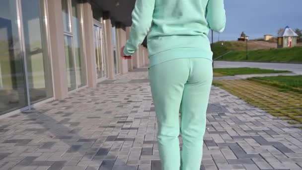 Een vrouw in een mint warme trainingspak loopt de straat op en danst. Roodharige meid in een blauwe sweater en broek kwispelt kont. Achteraanzicht. Een close-up. Vroeg zonnig ijzige ochtend. — Stockvideo