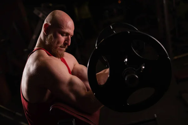 Egy szakállas kopasz ember bicepszeket gyakorol egy súlyzóval. A fiatalember testépítéssel foglalkozik. Edző az edzőteremben izmos karokkal — Stock Fotó