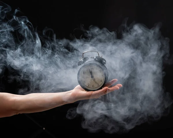 Una mujer sostiene un despertador en un estudio lleno de humo. Niebla blanca envolvió un reloj mecánico retro redondo . —  Fotos de Stock