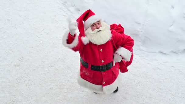 Visão superior sobre amigável Papai Noel ativamente gesticulando e desejando Feliz Natal. Um homem idoso em um traje de Papai Noel fica na neve do lado de fora e deseja um feliz ano novo. — Vídeo de Stock