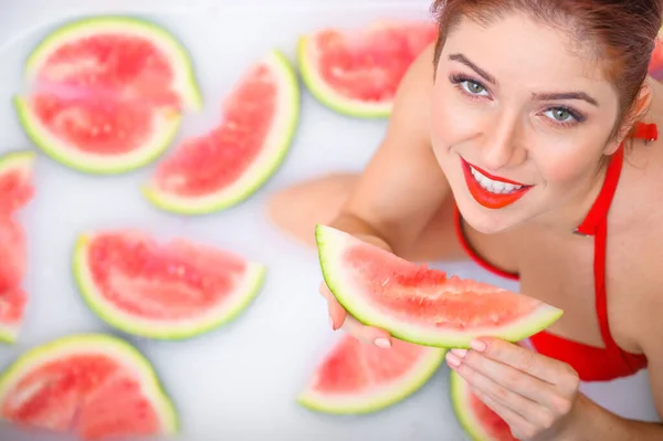 Portret van een vrouw in een rood badpak die een watermeloen eet en grijnst. Roodharig meisje met rode lippenstift neemt een bad met melk en fruit om haar huid te verjongen. Lichaamsverzorging. — Stockfoto