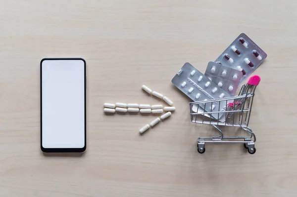 Online pharmacy. Blisters with a variety of pills in a mini shopping trolley and capsule arrow next to a white screen mobile phone. Smartphone app for buying medicine.