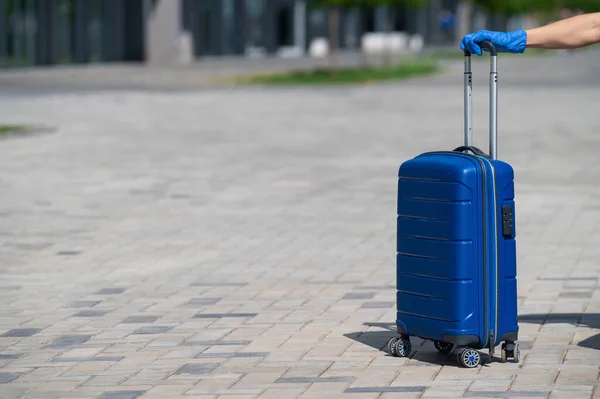 Gros plan d'une main féminine dans un gant sur un bagage. Une femme sans visage tient un grand sac bleu par la poignée coulissante dans la rue. Concept de voyage lors d'une épidémie de virus. Hygiène personnelle . — Photo