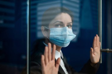 A sad quarantined woman in a business suit leaned against the window. Caucasian girl in a mask on isolation put her hands on a glass door. Maintaining social distance to prevent coronavirus. clipart
