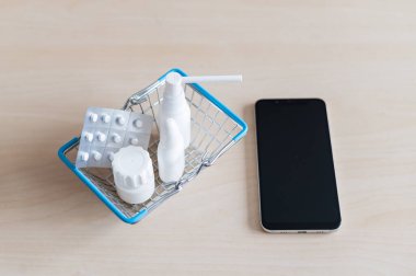 İnternetteki eczane. İçinde haplar olan su toplayıcı, siyah ekranlı cep telefonunun yanında küçük bir alışveriş sepetinde kapsüllü sprey. İlaç almak için akıllı telefon uygulaması.