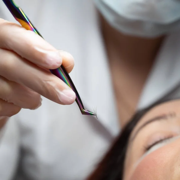 Donna si trova sulla procedura di estensione ciglia in un salone di bellezza. Lashmaker tiene pinzette con un mucchio di ciglia artificiali. Primo piano di un artigiano in guanti . — Foto Stock