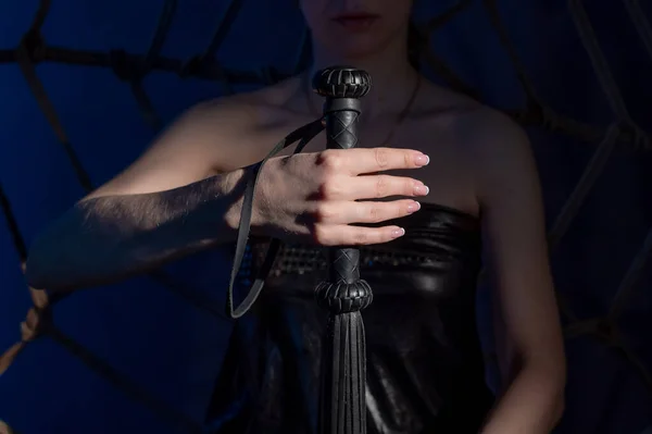 Концепція БДСМ. Жінка в шкіряній сукні тримає чорний батіг з довгою бахромою. Рольові ігри для дорослих. Крупним планом секс-іграшки для домінування . — стокове фото