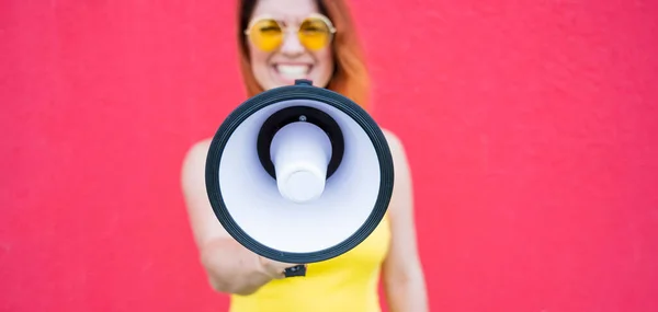 Eine Frau im Kleid mit Brille und Ohrringen steht im Profil vor gelbem Hintergrund und ruft in ein Megafon. Porträt eines Mädchens mit einem Lautsprecher. Lady schreit ein Schallverstärkungsgerät an. — Stockfoto