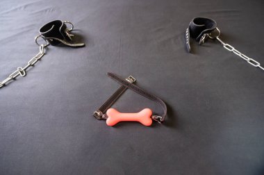 Kemik şeklinde bir şaka ve gri bir çarşafta zincirli deri kelepçeler. BDSM konsepti. Yetişkinler için rol yapma oyunları. Egemenlik ve itaat.