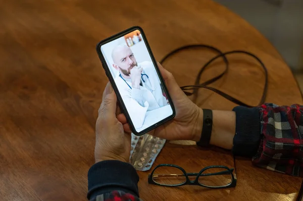 Онлайн медицина. Пожилая женщина с симптомами коронавируса разговаривает по смартфону с вирусологом. Мужчина-врач слушает жалобы на здоровье пожилой женщины на видео-ссылку . — стоковое фото
