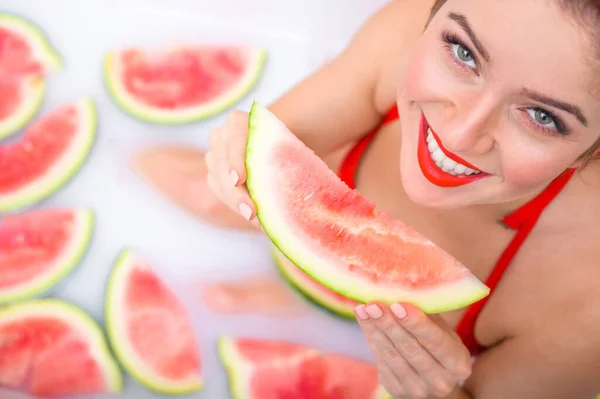 Portrait of a woman in a red swimsuit eating a watermelon and smirking. Redhead girl with red lipstick takes a bath with milk and fruit to rejuvenate her skin. Body care. — Stock Photo, Image