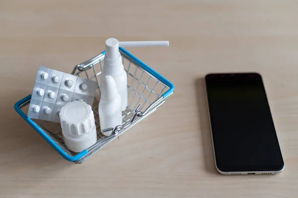 Online pharmacy. Blister with pills throat spray jar with capsules in a mini shopping basket next to a mobile phone with a black screen. Smartphone app for buying medicine.