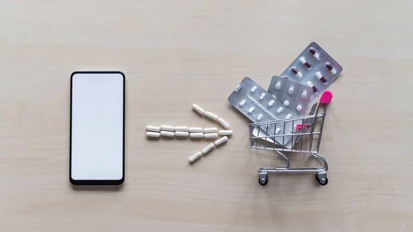 Online pharmacy. Blisters with a variety of pills in a mini shopping trolley and capsule arrow next to a white screen mobile phone. Smartphone app for buying medicine.