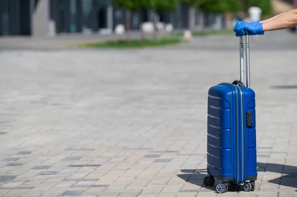 Nahaufnahme einer weiblichen Hand in einem Handschuh an einem Gepäckgriff. Eine Frau läuft die Straße hinunter und trägt eine große blaue Tasche. Reisekonzept während eines Virus-Ausbruchs. Körperpflege — Stockfoto