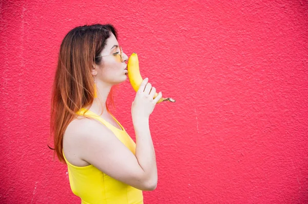 Woman in a yellow dress and glasses erotically kisses a banana on a red background. Portrait of a girl fantasizing about oral sex. Depicts Blowjob. Metaphor to the penis. — 스톡 사진