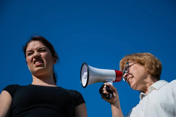 Конфлікт поколінь. Емоційна літня жінка кричить доньці в мегафоні. Літня мати клянеться жінкою середнього віку на гучномовному фоні . — стокове фото