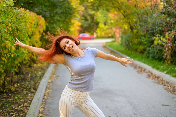 Eine schöne rothaarige Frau läuft die Straße entlang und tanzt. Glückliches Mädchen im grauen T-Shirt und gestreiften Leggings spaziert und hat Spaß im herbstlichen Park. Foto in Bewegung. — Stockfoto