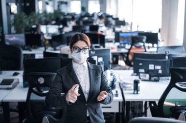 Tıbbi maske takmış kadın yönetici el antiseptiğine sprey sıkar. Takım elbiseli bir kadın dezenfekte için dezenfektan kullanıyor. Coronavirüs salgını sırasında ofis işi kavramı.