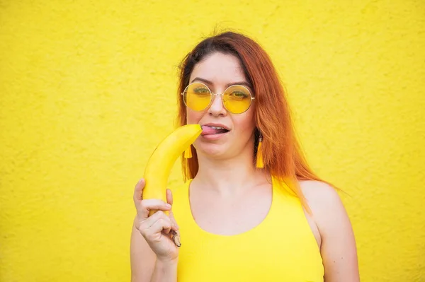 Красивая рыжеволосая женщина в жёлтом платье и солнечных очках показывает язык и облизывает банан. Портрет фантазирующей девушки с спелыми фруктами . — стоковое фото