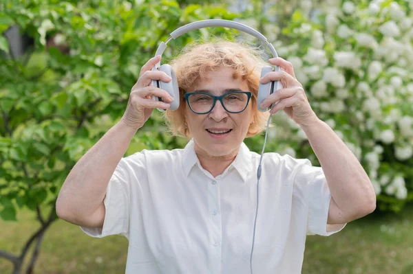 Glad gammal kvinna går i parken av blommande äppelträd och sätter på hörlurar. En kvinnlig pensionär njuter av en varm sommardag i trädgården och lyssnar på musik. Avancerad äldre generation. — Stockfoto