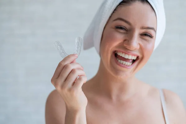 Uma mulher com uma toalha na cabeça segura um protetor bucal clareador para dentes. A menina alinha os dentes com a ajuda de retentores removíveis de noite transparentes. Dispositivo ortodôntico para um sorriso perfeito . — Fotografia de Stock