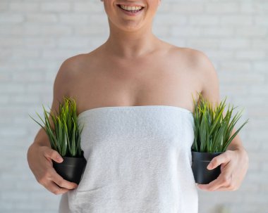 Beyaz havluyla yüzü olmayan kadın elinde bitkilerle iki kap tutuyor. Yeşil çalıların yardımıyla tanınmayan bir kız koltuk altı kıllarının bitki örtüsünü taklit ediyor. Depilasyon alanı. Kadın kılları alınıyor..