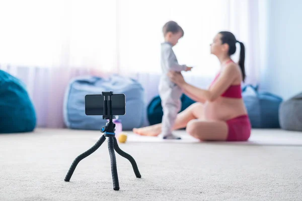 Une femme enceinte fait de la gymnastique avec son fils en direct. La future mère tourne une vidéo de formation pour son blog au téléphone. Fitness avec un bébé. — Photo