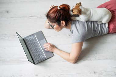 Kafkasyalı kızıl saçlı bir kadın küçük köpeğiyle yerde yatıyor ve dizüstü bilgisayarda yazıyor. Serbest çalışan bir kadın karantinada evden çalışıyor. Sadık köpek yavrusu Jack Russell Terrier.