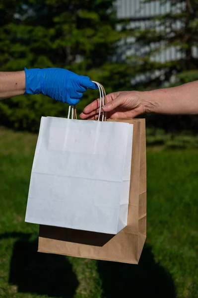 Um mensageiro sem rosto em luvas entrega um saco de papel ecológico a um cliente em um parque. O conceito de doação de produtos essenciais para idosos durante a epidemia do coronavírus . — Fotografia de Stock