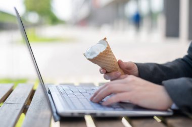 Yüzü olmayan bir kadın yazlık terasta bilgisayar başında çalışıyor ve vanilyalı dondurma yiyor. Dizüstü bilgisayarda yazı yazan ve elinde dondurma külahı tutan kadınların yakın çekimi. Yaz serinletici tatlı.